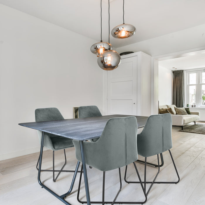 Esszimmer mit rechteckigem dunklem Holztisch und vier grauen Samtstühlen mit Metallgestell, darüber drei runde Hängelampen mit warmem Licht