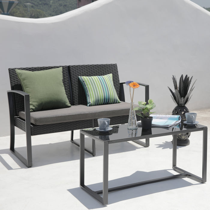 Schwarzes Rattan-Garten-Sofa mit grauem Sitzkissen und zwei Kissen in Grün- und Blautönen, davor ein schwarzer Glastisch mit zwei Tassen, einer Vase mit Blumen und einer Pflanze, weißer Hintergrund