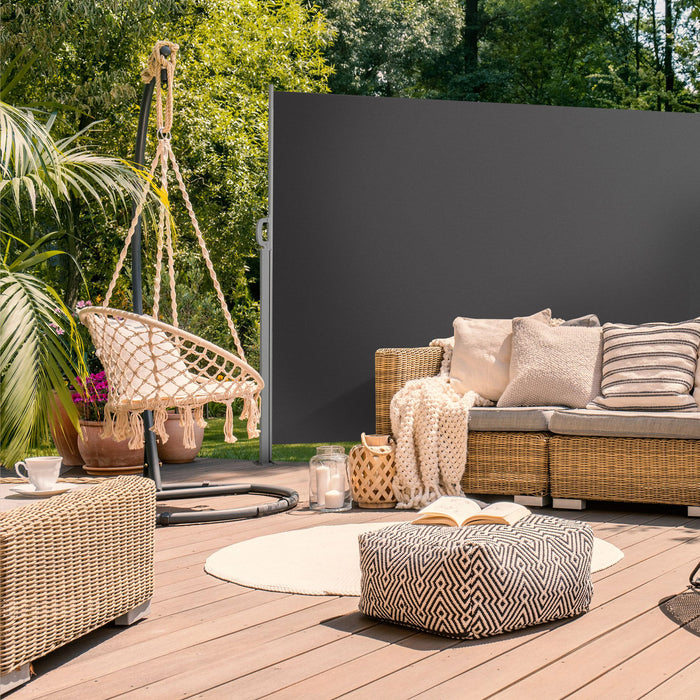 Gemütliche Terrasse mit Rattansofa, Hängesessel, Kissen, Decke und rundem Teppich, umgeben von grünen Pflanzen und mit dunkelgrauer Seitenmarkise geschützt