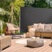Gemütliche Terrasse mit Rattansofa, Hängesessel, Kissen, Decke und rundem Teppich, umgeben von grünen Pflanzen und mit dunkelgrauer Seitenmarkise geschützt