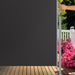 Hand hält eine dunkelgraue Sichtschutzwand auf einer Holzterrasse mit pinken Blumen und grünen Büschen im Hintergrund