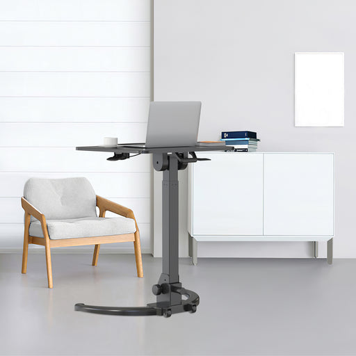 Stehschreibtisch mit Laptop und Tasse neben einem grauen Sessel mit Holzrahmen in einem hellen Raum mit weißem Sideboard und leerem Bilderrahmen