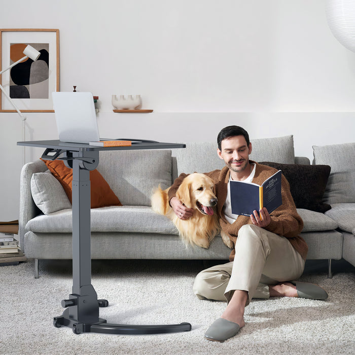 Mann sitzt auf dem Boden vor einem grauen Sofa, liest ein Buch und streichelt einen Golden Retriever, neben ihm ein höhenverstellbarer Tisch mit Laptop