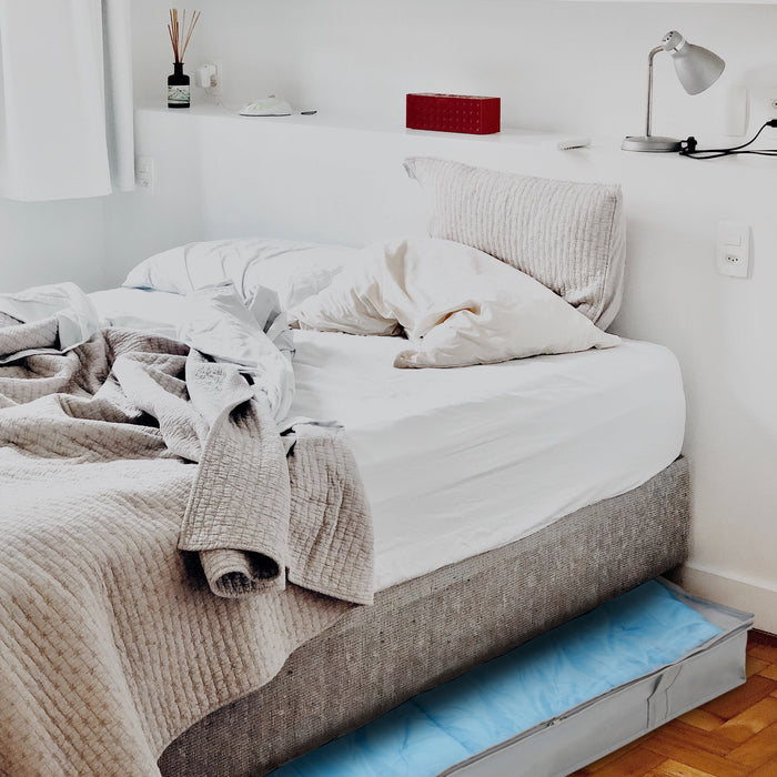 Eine graue Aufbewahrungstasche schaut unter einem Bett mit weißen und grauen Bettwäsche hervor