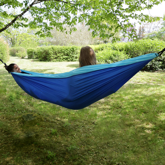 Zwei Personen liegen in einer blauen Hängematte unter einem Baum in einem grünen Garten