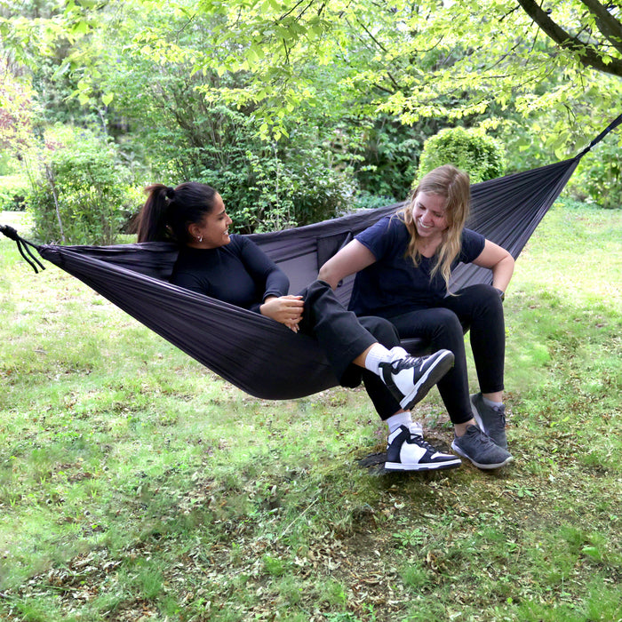 Zwei Personen sitzen in einer grauen Hängematte unter einem Baum in einem grünen Garten