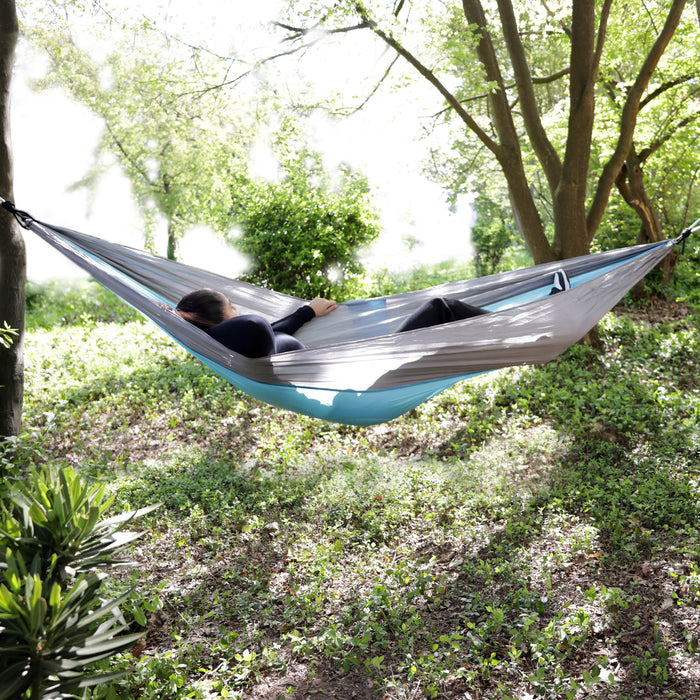 Person liegt entspannt in einer blauen und grauen Hängematte zwischen Bäumen im grünen Garten
