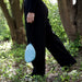 Person in schwarzer Hose und schwarzen Schuhen geht durch einen bewachsenen Wald und trägt eine kleine blaue Tasche mit Hängematte darin in der Hand