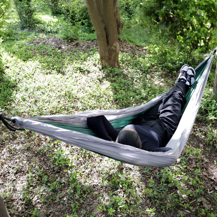 Person liegt entspannt in einer Hängematte zwischen Bäumen im Wald, trägt schwarze Kleidung und weiße Sneakers