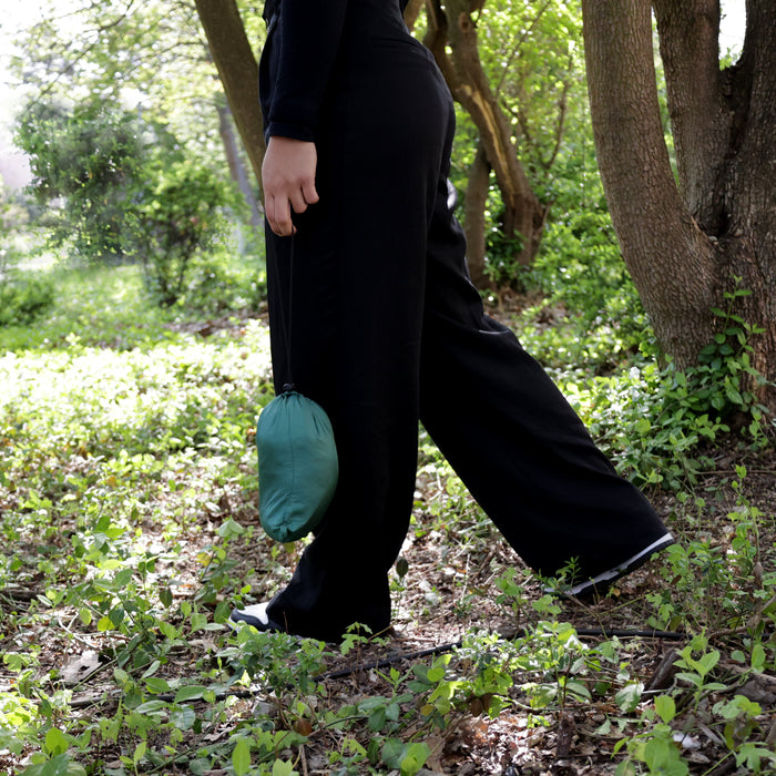 Person in schwarzer Hose und schwarzen Schuhen geht durch einen bewachsenen Wald und trägt eine kleine grüne Tasche mit Hängematte darin in der Hand