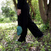 Person in schwarzer Hose und schwarzen Schuhen geht durch einen bewachsenen Wald und trägt eine kleine grüne Tasche mit Hängematte darin in der Hand