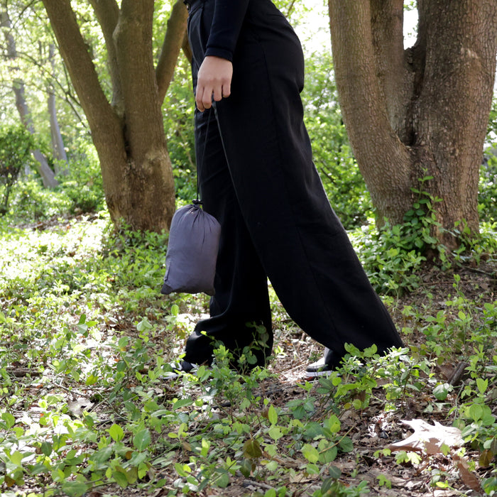 Person in schwarzer Hose und schwarzen Schuhen geht durch einen bewachsenen Wald und trägt eine kleine graue Tasche mit Hängematte darin in der Hand