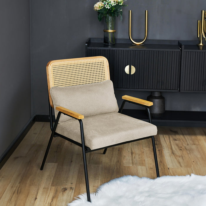 Cremefarbener Loungestuhl mit Rattan-Rückenlehne und Holzarmlehnen vor schwarzem Sideboard mit goldfarbenen Griffen