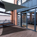 Große Terrasse mit dunklem Holzboden, Glas-Schiebetüren und grauem Sonnensegel 3,6x3,6x5,1m an modernem Gebäude mit Backstein und blauen Fassadenelementen