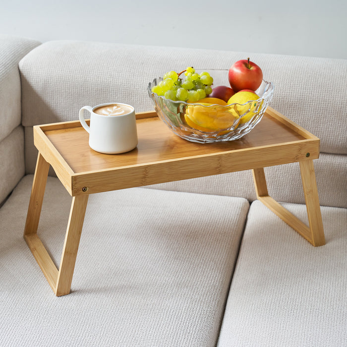 Bambus-Tablett mit klappbaren Beinen auf einem hellen Sofa, darauf eine Glasschale mit verschiedenen Früchten und eine Tasse Kaffee mit Milchschaum