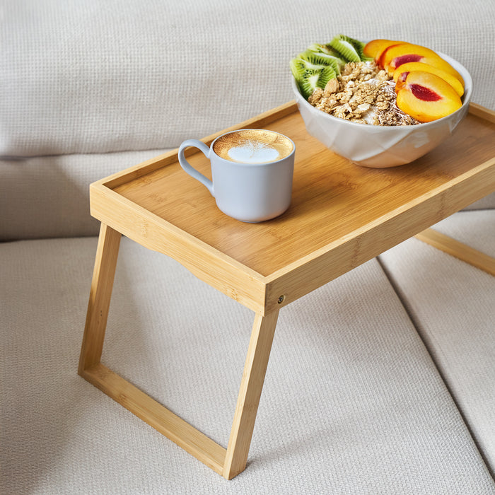 Bambus-Tablett mit klappbaren Beinen auf einem hellen Sofa, darauf eine Glasschale mit Müsli und eine Tasse Kaffee mit Milchschaum