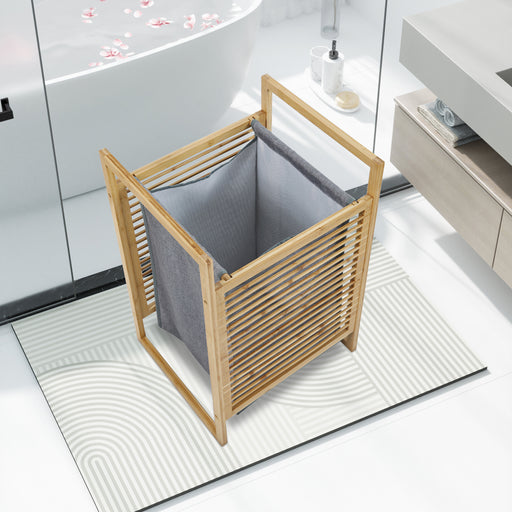 Moderner Wäschekorb aus Holz mit grauem Stoffeinsatz steht auf einem weißen Badezimmerteppich neben Badewanne und Waschbecken