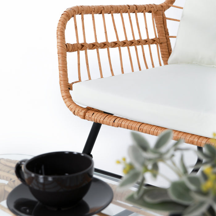 Rattanbank mit weißem Sitzkissen und schwarzer Tasse auf Untertasse auf einem Glastisch, unscharfe grüne Pflanze im Vordergrund