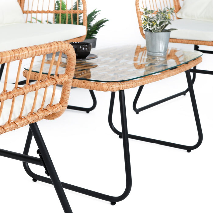 Glas-Couchtisch mit schwarzem Metallgestell zwischen zwei Rattanstühlen mit weißen Kissen, Pflanze in Metalltopf auf dem Tisch